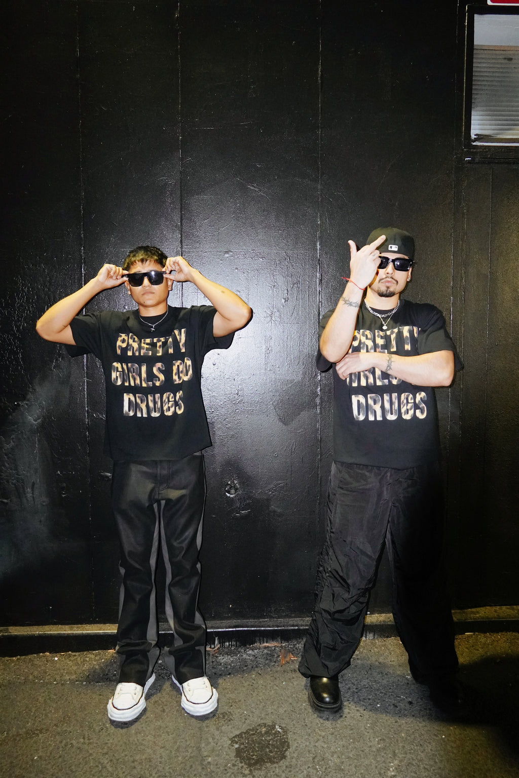 Two people wearing black t-shirts with text, posing in front of a dark wall.
