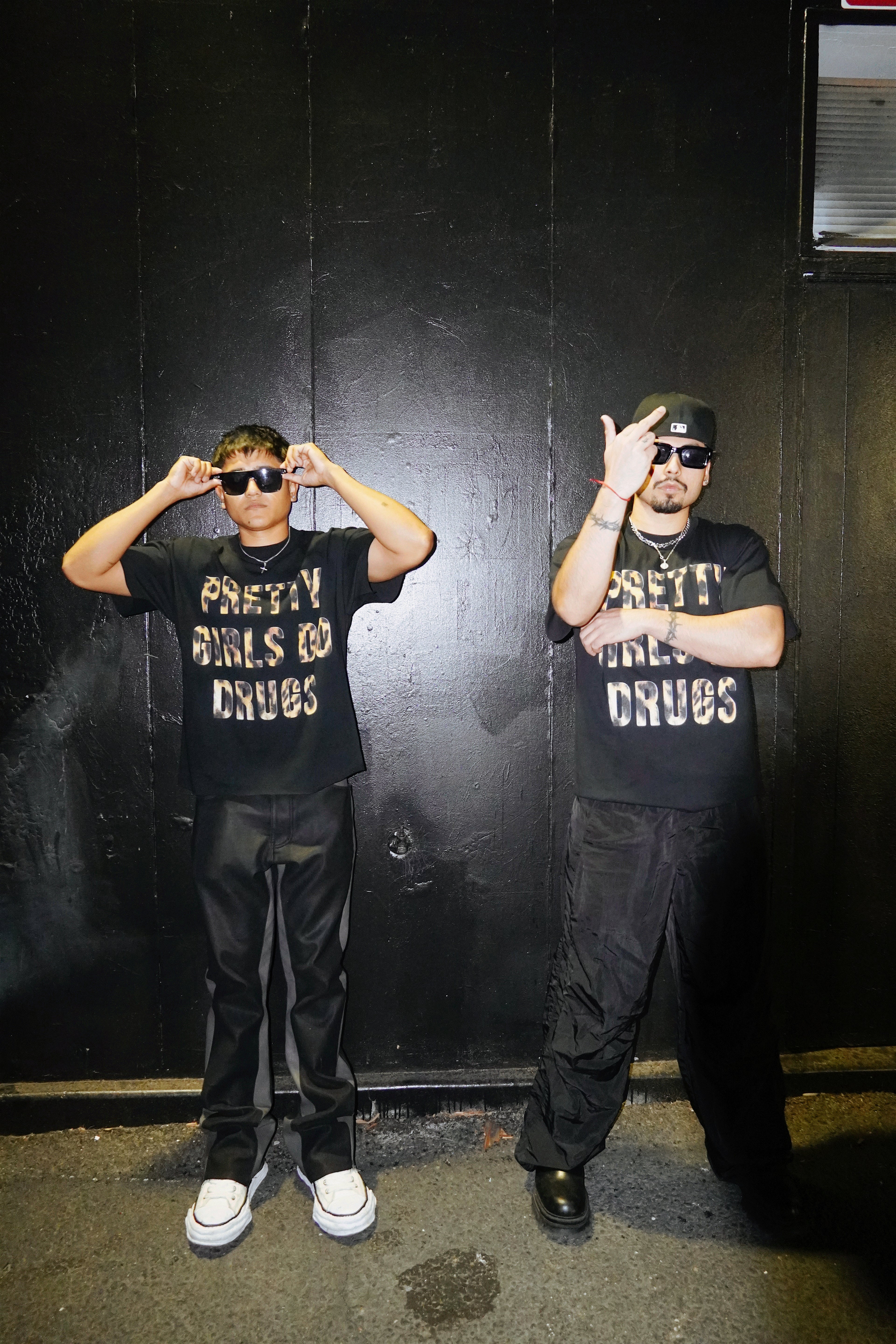 Two people wearing black t-shirts with text, posing in front of a dark wall.
